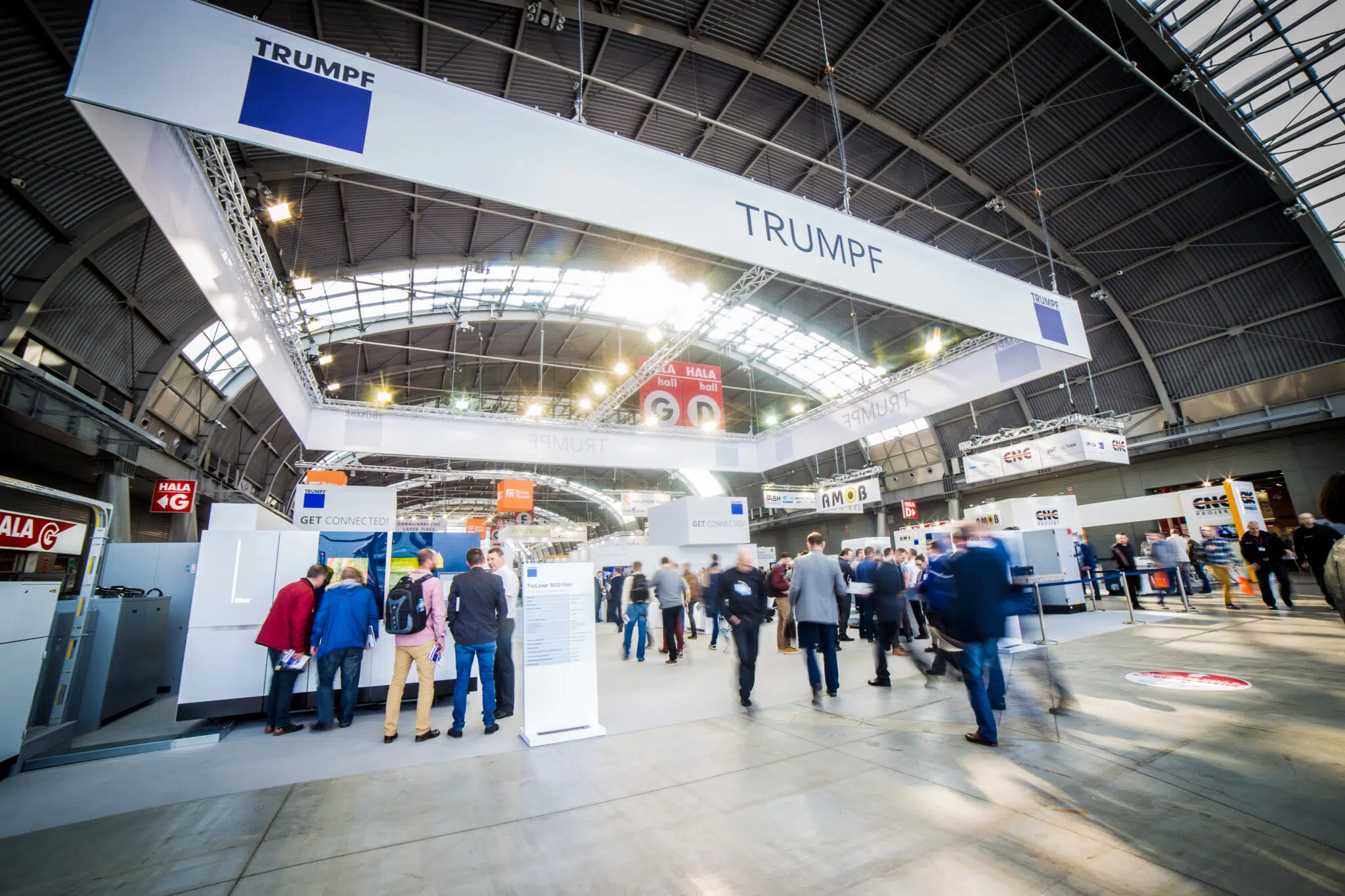 Na zdjęciach widzimy stoisko targowe zwane ekspozycję targową marki TRUMPF na targach STOM Kielce z imponującymi podwieszeniami sufitowymi, które budują nowoczesny klimat stoiska. Przestrzeń została zaprojektowana w sposób otwarty, łącząc strefy prezentacji maszyn z eleganckimi miejscami do rozmów biznesowych. Całość podkreśla technologiczny charakter marki poprzez surowe, industrialne detale i precyzyjnie rozmieszczone oświetlenie punktowe.