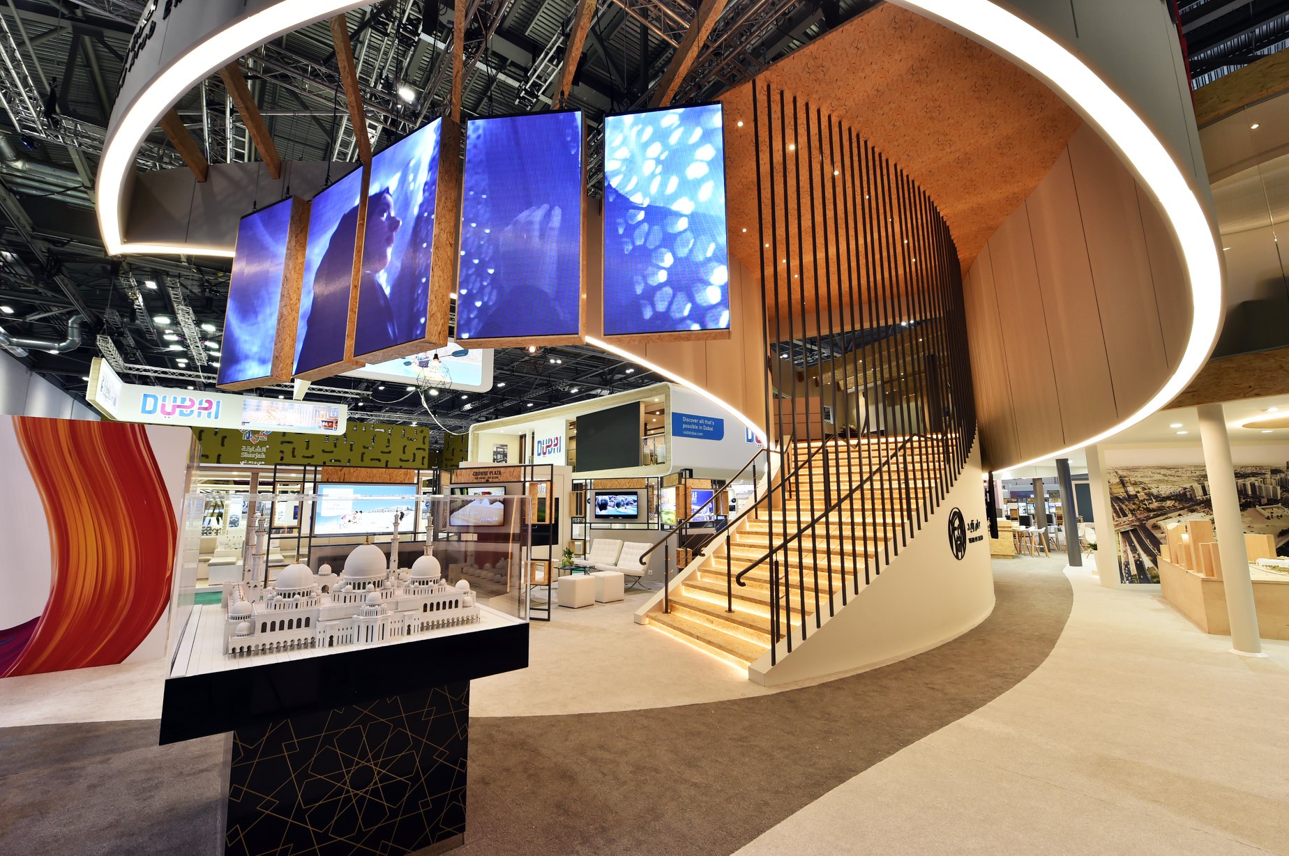 Na zdjęciach widzimy stoisko targowe marki Abu Dhabi na targach WTM London w 2018 roku o powierzchni 700 m2 parter i 170 m2 piętro. Prezentowana realizacja to zaawansowany konstrukcyjnie projekt stoiska piętrowego, łączący nowoczesną architekturę targową z funkcjonalnością stref premium. Jako eksperci w budowie stoisk targowych, postawiliśmy na synergię naturalnych materiałów i technologii multimedialnych. Konstrukcja Nośna: Wytrzymały system stalowy umożliwiający budowę bezpiecznego piętra, wykończony estetyczną zabudową z płyt MDF oraz elementami z naturalnego drewna (sklejka, płyty OSB w designerskim wydaniu). Schody i Komunikacja: Reprezentacyjne, szerokie schody zintegrowane z bryłą stoiska, wyposażone w liniowe podświetlenie LED stopni, zapewniające bezpieczeństwo i prestiżowy wygląd. Multimadia i Oświetlenie: Centralnym punktem stoiska jest podwieszany pierścień z wielkoformatowymi ekranami LED, które gwarantują widoczność marki z najdalszych punktów hali. Oświetlenie architektoniczne oparte na profilach LED podkreśla organiczne kształty zabudowy. Detale i Wykończenie: Ażurowe ścianki z pionowych lameli oddzielają strefę biznesową, zapewniając intymność rozmów przy jednoczesnym zachowaniu lekkości wizualnej. Na parterze wydzielono strefę ekspozycyjną z makietami i interaktywnymi panelami.