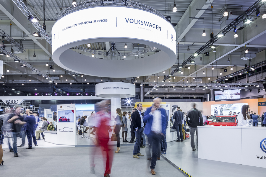 Realizacja zabudowy dla wiodących marek motoryzacyjnych podczas Poznań Motor Show to monumentalny projekt o łącznej powierzchni przekraczającej 4500 m². Jako BWS EXPO zapewniliśmy kompleksowe wsparcie techniczne i wykonawcze, tworząc spójne, a jednocześnie unikalne przestrzenie ekspozycyjne dla marek takich jak BMW, Porsche, Audi, Rolls-Royce oraz całej grupy Volkswagen (Skoda, Seat, VW). Kluczowe aspekty techniczno-wykonawcze: Skala i Logistyka: Równoczesna budowa stoisk dla wielu marek premium wymagała perfekcyjnej koordynacji prac montażowych oraz logistyki materiałowej w kilku halach targowych. Design i Materiały: Każda przestrzeń została wykończona zgodnie z rygorystycznymi standardami wizualnymi (Corporate Identity) poszczególnych brandów. Wykorzystano m.in. eleganckie okładziny drewniane dla stref luksusowych (Rolls-Royce, Porsche), nowoczesne konstrukcje loftowe oraz wysokiej jakości posadzki targowe. Systemy Podwieszeń i Multimeadia: Zastosowano zaawansowane konstrukcje kratownicowe z wielkoformatowymi ekranami LED, dynamicznym oświetleniem scenicznym oraz potężnymi elementami brandingowymi (np. charakterystyczne ringi Volkswagen Financial Services czy kubiki BMW), które zapewniały widoczność marek z dużej odległości. Strefy Specjalne: Realizacja objęła budowę dwupoziomowych stref VIP, butików Lifestyle Shop (BMW), profesjonalnych recepcji z grafikami oraz interaktywnych stoisk usług finansowych. Dzięki doświadczeniu w obsłudze największych wydarzeń motoryzacyjnych, dostarczyliśmy infrastrukturę, która połączyła technologiczną nowoczesność z prestiżem, tworząc idealne tło dla premier motoryzacyjnych najwyższej klasy.