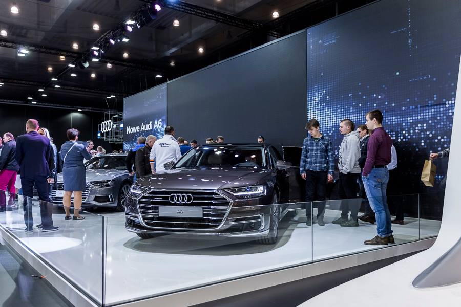 Realizacja zabudowy dla wiodących marek motoryzacyjnych podczas Poznań Motor Show to monumentalny projekt o łącznej powierzchni przekraczającej 4500 m². Jako BWS EXPO zapewniliśmy kompleksowe wsparcie techniczne i wykonawcze, tworząc spójne, a jednocześnie unikalne przestrzenie ekspozycyjne dla marek takich jak BMW, Porsche, Audi, Rolls-Royce oraz całej grupy Volkswagen (Skoda, Seat, VW). Kluczowe aspekty techniczno-wykonawcze: Skala i Logistyka: Równoczesna budowa stoisk dla wielu marek premium wymagała perfekcyjnej koordynacji prac montażowych oraz logistyki materiałowej w kilku halach targowych. Design i Materiały: Każda przestrzeń została wykończona zgodnie z rygorystycznymi standardami wizualnymi (Corporate Identity) poszczególnych brandów. Wykorzystano m.in. eleganckie okładziny drewniane dla stref luksusowych (Rolls-Royce, Porsche), nowoczesne konstrukcje loftowe oraz wysokiej jakości posadzki targowe. Systemy Podwieszeń i Multimeadia: Zastosowano zaawansowane konstrukcje kratownicowe z wielkoformatowymi ekranami LED, dynamicznym oświetleniem scenicznym oraz potężnymi elementami brandingowymi (np. charakterystyczne ringi Volkswagen Financial Services czy kubiki BMW), które zapewniały widoczność marek z dużej odległości. Strefy Specjalne: Realizacja objęła budowę dwupoziomowych stref VIP, butików Lifestyle Shop (BMW), profesjonalnych recepcji z grafikami oraz interaktywnych stoisk usług finansowych. Dzięki doświadczeniu w obsłudze największych wydarzeń motoryzacyjnych, dostarczyliśmy infrastrukturę, która połączyła technologiczną nowoczesność z prestiżem, tworząc idealne tło dla premier motoryzacyjnych najwyższej klasy.