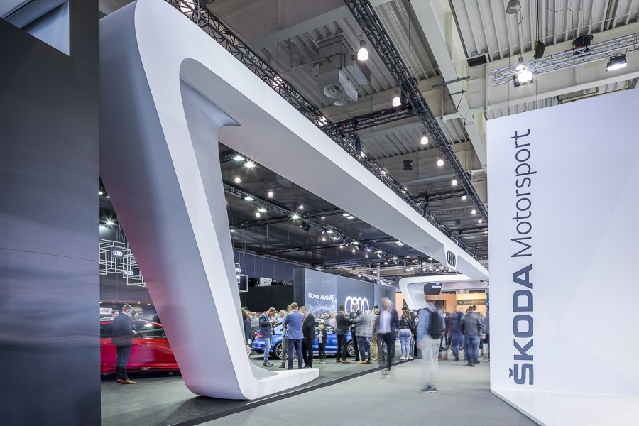 Realizacja zabudowy dla wiodących marek motoryzacyjnych podczas Poznań Motor Show to monumentalny projekt o łącznej powierzchni przekraczającej 4500 m². Jako BWS EXPO zapewniliśmy kompleksowe wsparcie techniczne i wykonawcze, tworząc spójne, a jednocześnie unikalne przestrzenie ekspozycyjne dla marek takich jak BMW, Porsche, Audi, Rolls-Royce oraz całej grupy Volkswagen (Skoda, Seat, VW). Kluczowe aspekty techniczno-wykonawcze: Skala i Logistyka: Równoczesna budowa stoisk dla wielu marek premium wymagała perfekcyjnej koordynacji prac montażowych oraz logistyki materiałowej w kilku halach targowych. Design i Materiały: Każda przestrzeń została wykończona zgodnie z rygorystycznymi standardami wizualnymi (Corporate Identity) poszczególnych brandów. Wykorzystano m.in. eleganckie okładziny drewniane dla stref luksusowych (Rolls-Royce, Porsche), nowoczesne konstrukcje loftowe oraz wysokiej jakości posadzki targowe. Systemy Podwieszeń i Multimeadia: Zastosowano zaawansowane konstrukcje kratownicowe z wielkoformatowymi ekranami LED, dynamicznym oświetleniem scenicznym oraz potężnymi elementami brandingowymi (np. charakterystyczne ringi Volkswagen Financial Services czy kubiki BMW), które zapewniały widoczność marek z dużej odległości. Strefy Specjalne: Realizacja objęła budowę dwupoziomowych stref VIP, butików Lifestyle Shop (BMW), profesjonalnych recepcji z grafikami oraz interaktywnych stoisk usług finansowych. Dzięki doświadczeniu w obsłudze największych wydarzeń motoryzacyjnych, dostarczyliśmy infrastrukturę, która połączyła technologiczną nowoczesność z prestiżem, tworząc idealne tło dla premier motoryzacyjnych najwyższej klasy.