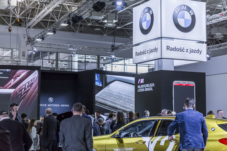 Realizacja zabudowy dla wiodących marek motoryzacyjnych podczas Poznań Motor Show to monumentalny projekt o łącznej powierzchni przekraczającej 4500 m². Jako BWS EXPO zapewniliśmy kompleksowe wsparcie techniczne i wykonawcze, tworząc spójne, a jednocześnie unikalne przestrzenie ekspozycyjne dla marek takich jak BMW, Porsche, Audi, Rolls-Royce oraz całej grupy Volkswagen (Skoda, Seat, VW). Kluczowe aspekty techniczno-wykonawcze: Skala i Logistyka: Równoczesna budowa stoisk dla wielu marek premium wymagała perfekcyjnej koordynacji prac montażowych oraz logistyki materiałowej w kilku halach targowych. Design i Materiały: Każda przestrzeń została wykończona zgodnie z rygorystycznymi standardami wizualnymi (Corporate Identity) poszczególnych brandów. Wykorzystano m.in. eleganckie okładziny drewniane dla stref luksusowych (Rolls-Royce, Porsche), nowoczesne konstrukcje loftowe oraz wysokiej jakości posadzki targowe. Systemy Podwieszeń i Multimeadia: Zastosowano zaawansowane konstrukcje kratownicowe z wielkoformatowymi ekranami LED, dynamicznym oświetleniem scenicznym oraz potężnymi elementami brandingowymi (np. charakterystyczne ringi Volkswagen Financial Services czy kubiki BMW), które zapewniały widoczność marek z dużej odległości. Strefy Specjalne: Realizacja objęła budowę dwupoziomowych stref VIP, butików Lifestyle Shop (BMW), profesjonalnych recepcji z grafikami oraz interaktywnych stoisk usług finansowych. Dzięki doświadczeniu w obsłudze największych wydarzeń motoryzacyjnych, dostarczyliśmy infrastrukturę, która połączyła technologiczną nowoczesność z prestiżem, tworząc idealne tło dla premier motoryzacyjnych najwyższej klasy.