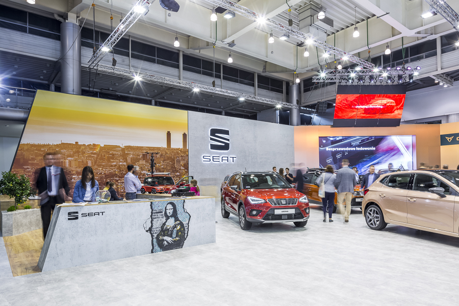 Realizacja zabudowy dla wiodących marek motoryzacyjnych podczas Poznań Motor Show to monumentalny projekt o łącznej powierzchni przekraczającej 4500 m². Jako BWS EXPO zapewniliśmy kompleksowe wsparcie techniczne i wykonawcze, tworząc spójne, a jednocześnie unikalne przestrzenie ekspozycyjne dla marek takich jak BMW, Porsche, Audi, Rolls-Royce oraz całej grupy Volkswagen (Skoda, Seat, VW). Kluczowe aspekty techniczno-wykonawcze: Skala i Logistyka: Równoczesna budowa stoisk dla wielu marek premium wymagała perfekcyjnej koordynacji prac montażowych oraz logistyki materiałowej w kilku halach targowych. Design i Materiały: Każda przestrzeń została wykończona zgodnie z rygorystycznymi standardami wizualnymi (Corporate Identity) poszczególnych brandów. Wykorzystano m.in. eleganckie okładziny drewniane dla stref luksusowych (Rolls-Royce, Porsche), nowoczesne konstrukcje loftowe oraz wysokiej jakości posadzki targowe. Systemy Podwieszeń i Multimeadia: Zastosowano zaawansowane konstrukcje kratownicowe z wielkoformatowymi ekranami LED, dynamicznym oświetleniem scenicznym oraz potężnymi elementami brandingowymi (np. charakterystyczne ringi Volkswagen Financial Services czy kubiki BMW), które zapewniały widoczność marek z dużej odległości. Strefy Specjalne: Realizacja objęła budowę dwupoziomowych stref VIP, butików Lifestyle Shop (BMW), profesjonalnych recepcji z grafikami oraz interaktywnych stoisk usług finansowych. Dzięki doświadczeniu w obsłudze największych wydarzeń motoryzacyjnych, dostarczyliśmy infrastrukturę, która połączyła technologiczną nowoczesność z prestiżem, tworząc idealne tło dla premier motoryzacyjnych najwyższej klasy.